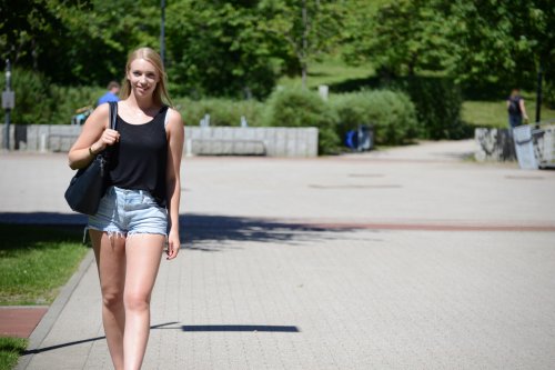 Studentin Paula läuft an einem Sommertag über den Campus. Im Hintergrund ein gepflasterter Weg und Grünflächen und Bäume.