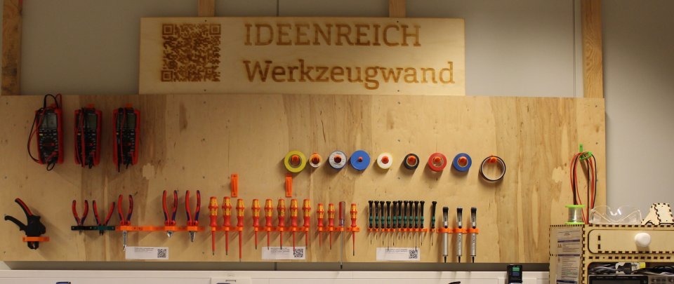 Holzplatte, an der viele Werkzeuge hängen, an einer Wand. Darüber ein Schild "IDEENREICH Werkzeugwand".