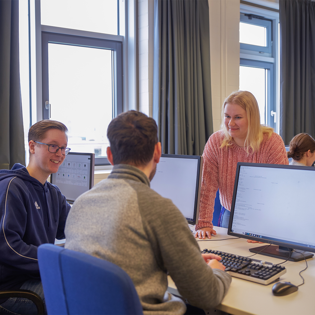 Studierende in einem Seminarraum. Zwei Studenten schauen auf Bildschirme, eine Studentin steht dahinter. Sie unterhalten sich.