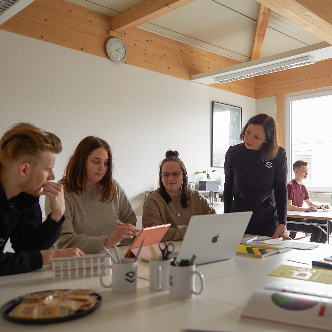 Studierende arbeiten in einem hellen, freundlichen Raum an Laptops. Eine DSozentin steht daneben und hört interessiert zu.