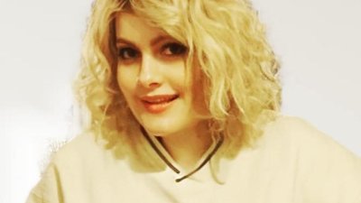 Eine junge Frau mit lockigen, blonden Haaren blickt selbstbewusst in die Kamera. Sie trägt einen weißen Pullover mit dem Aufdruck "Chicago" in Großbuchstaben.