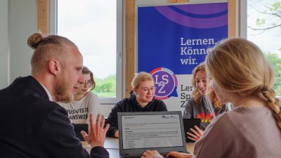 Einige Studierende sitzen am Tisch und lernen. Im Hintergrund sieht man das Logo des Lern- und Sprachenzentrums.