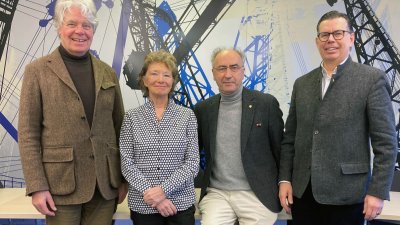 Vier Menschen, drei davon Männer, eine Frau, posieren für ein Gruppenbild und lächeln in die Kamera. Sie lehnen oder sitzen auf einem Tisch.