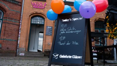 Aufstell-Tafel vor einem Lokal. Darauf Hinweise auf zwei Vorträge, an der Tafel sind bunte Luftballons festgebunden.