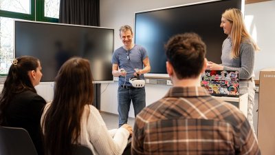 Ein lachender Mann mit VR Brille in der Hand steht vor Studierenden