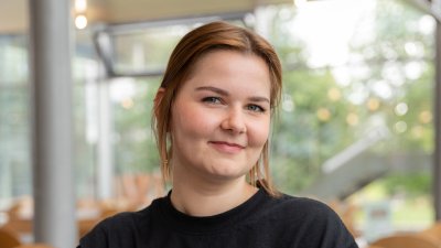 Junge Frau in schwarzem Oberteil sitzt in der Flensburger Mensa an einem Laptop und schaut in die Kamera.