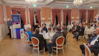 Ein Saal mit runden Tischen an denen Menschen sitzen, die einem Mann zuhören, der an einem Rednerpult steht und spricht