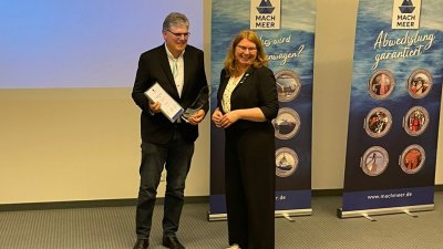 Ein Mann und eine Frau stehen auf einer Bühne. Sie übergibt ihm eine Urkunde