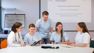 fünf Menschen sitzen am Tisch, einer steht, sie unterhalten sich, haben weiße Kittel an