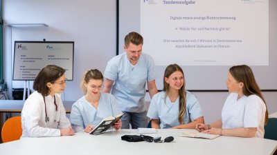 fünf Menschen sitzen am Tisch, einer steht, sie unterhalten sich, haben weiße Kittel an