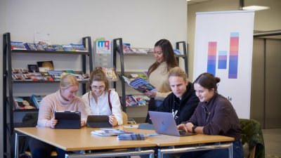 Fünf Studierende arbeiten gemeinsam an einem Tisch mit Laptop, Tablet und Broschüren. Die Szene zeigt, wie Lernen an der Hochschule zunehmend digital, kollaborativ und medienübergreifend stattfindet.