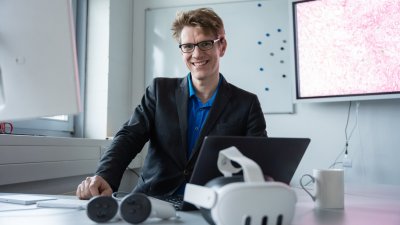 Person an einem Arbeitsplatz mit Laptop und VR-Brillen; im Hintergrund zeigt ein Bildschirm eine stark vergrößerte medizinische Gewebeaufnahme.