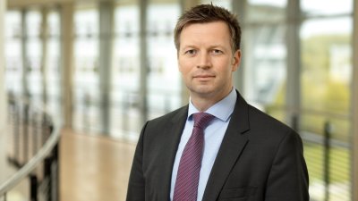 Porträt von Prof. Dr. Ilja Tuschy im Anzug vor einer hellen Fensterfront im Gebäude der Hochschule Flensburg.