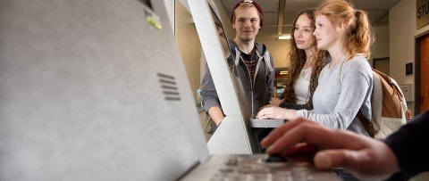 Drei Studierende an einem Infoterminal im Hauptgebäude. Vorn im Bild ist unscharf eine Hand auf der Tastatur eines weiteren Terminals zu erkennen.