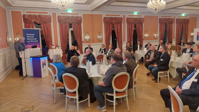 Ein Saal mit runden Tischen an denen Menschen sitzen, die einem Mann zuhören, der an einem Rednerpult steht und spricht