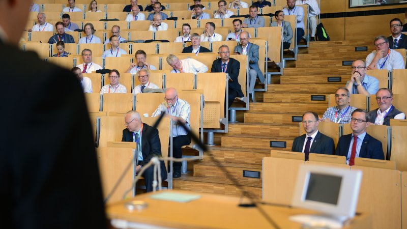 Blick in den Zuschauerraum des Audimax, Tagungsgäste schauen nach vorn. Vorne im Bild sieht man die Schulter eines Redners im Anzug.