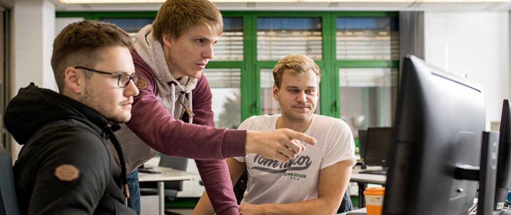 Drei Studenten sitzen in einem Computerlabor gemeinsam vor einem PC. Einer von ihnen zeigt mit dem Finger auf den Bildschirm.