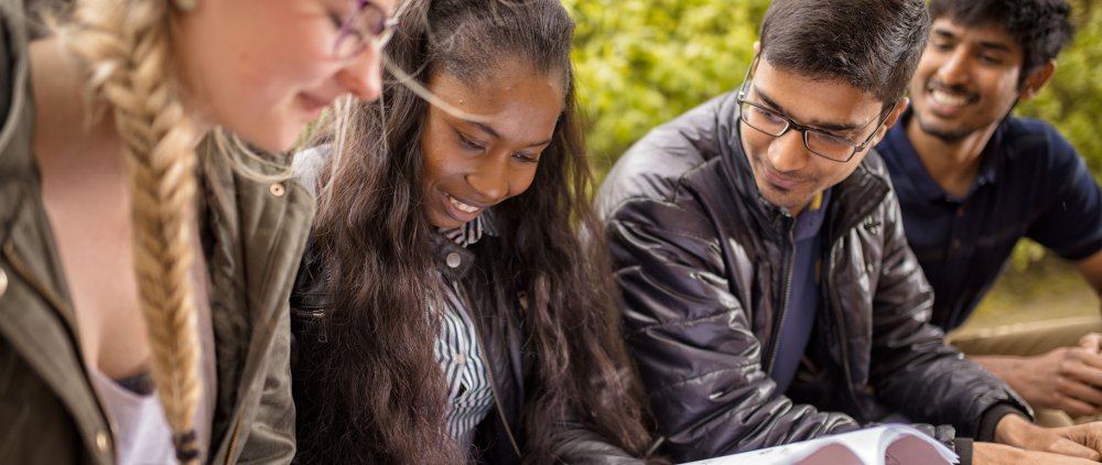 Vier Studierende, die ihrem Aussehen nach zu urteilen unterschiedliche kulturelle Hintergründe haben, sehen sich auf dem Campus sitzend gemeinsam etwas an, das nicht im Bild ist.