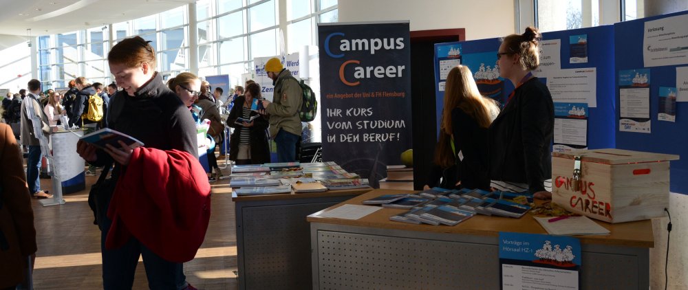 Messesituation: rechts vorn im Bild der Messestand des CampusCareer mit Postern, Broschüren usw. Links etwas abseits zwei Studentinnen, die in Broschüren blättern. Im Hintergrund Menschen und andere Messestände.