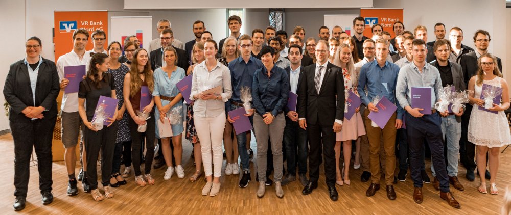 Gruppenbild aller Absolventinnen und Absolventen zusammen mit Lehrenden vor Aufstellern der VR Bank und der Hochschule.