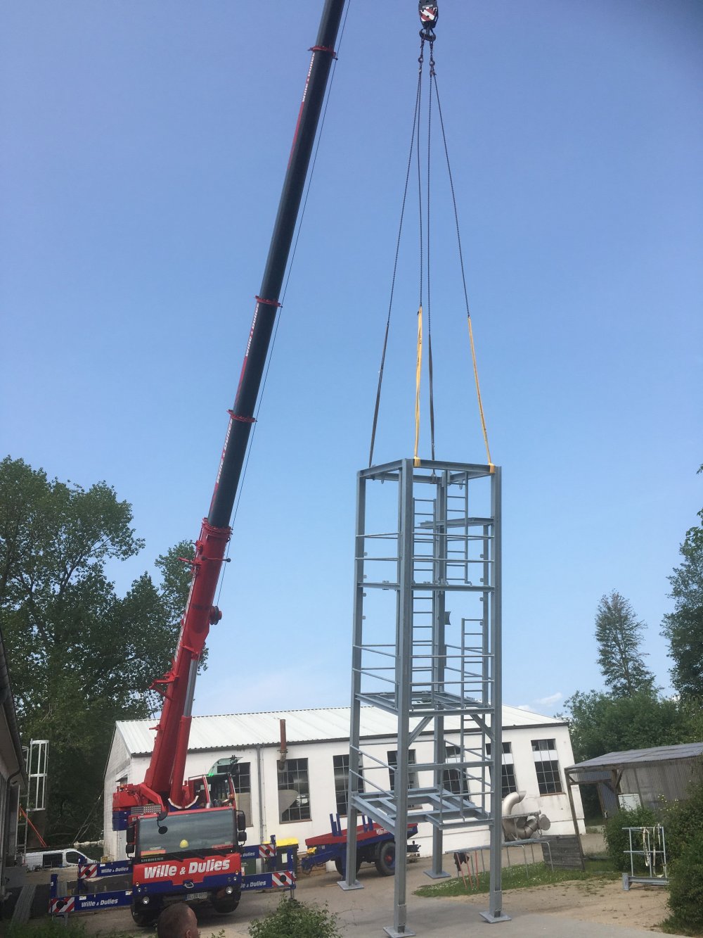 Vor blauem Himmel steht ein Kran, der mithilfe von Drahtseilen ein Metallgerüst aufrecht hält. Das Gerüst überragt das eingeschossige Gebäude im Hintergrund deutlich.