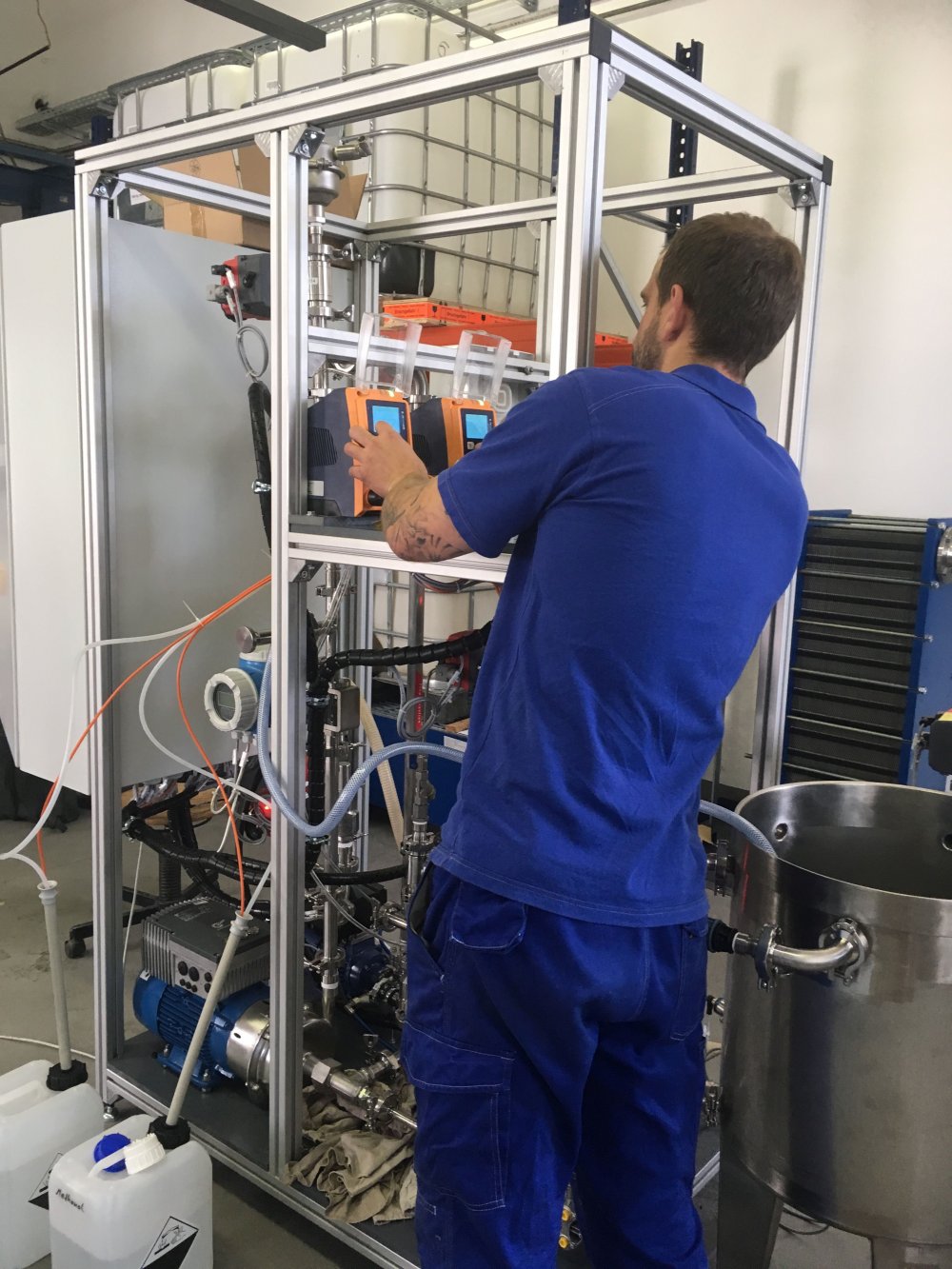 Ein Mann in dunkelblauen Arbeitshosen und gleichfarbigem T-Shirt steht vor einer Anlage, die aus Metallstreben gebildet wird. In der Anlage sind Kabel und Rohre zu sehen, außerdem zwei Eingabegeräte. Eines davon bedient er.