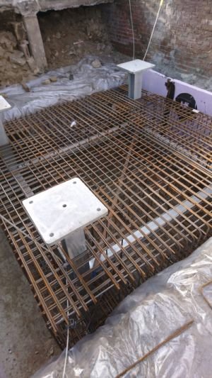 Ein Gitter aus Metall. Darum ist Plastikfolie und lose Steine und Reste von Bauschutt zu sehen, direkt an eine Seite angrenzend eine Mauer.