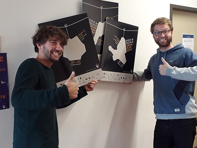 Zwei Studenten posieren mit &quot;Daumen hoch&quot;-Geste vor den Postern.