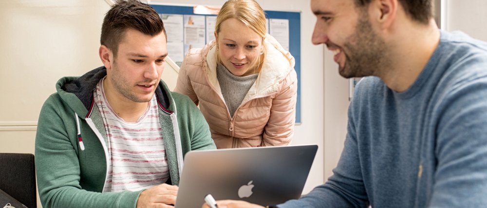 3 junge Menschen sind um einen Laptop versammelt und schauen interessiert auf dessen Bildschirm.