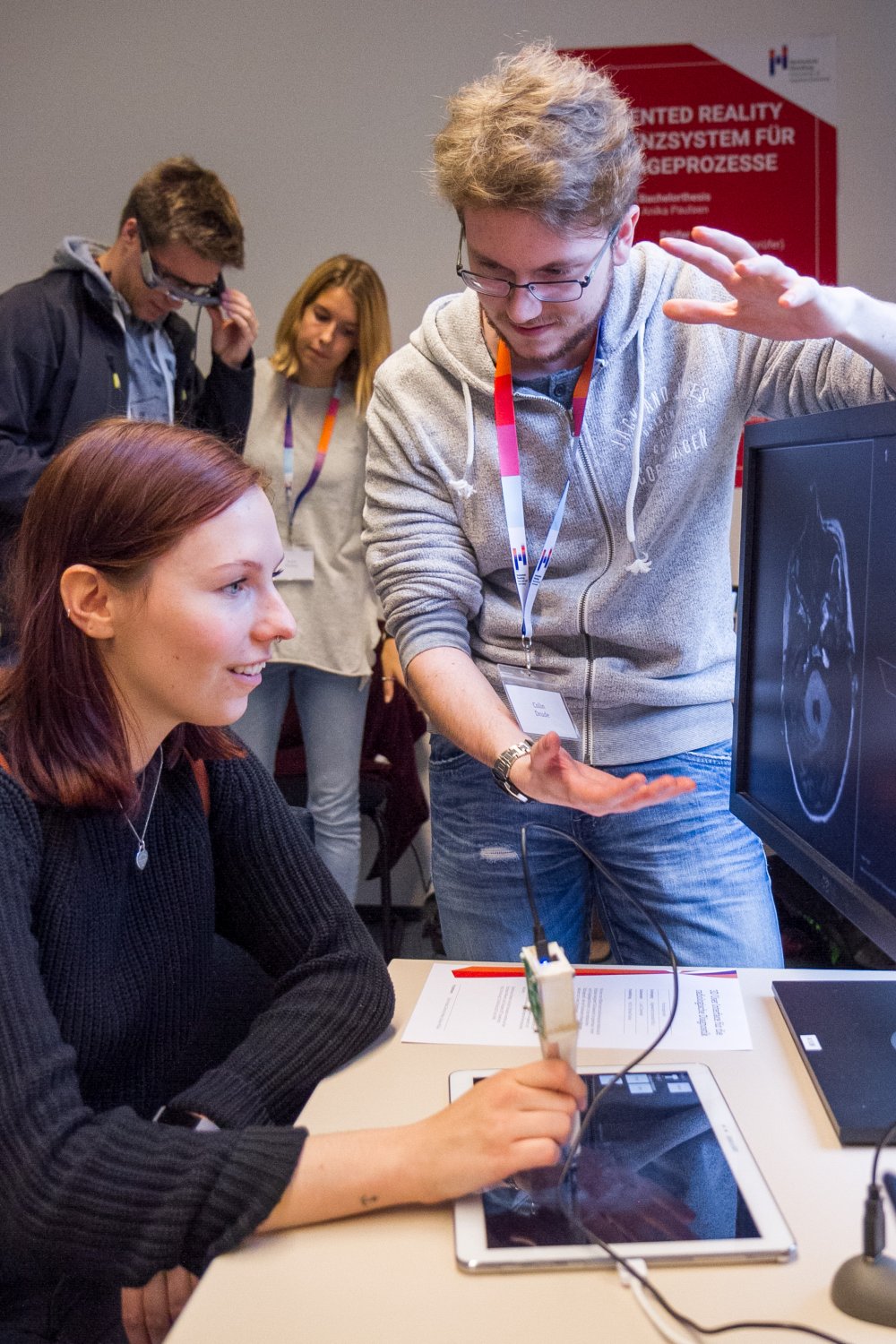 Sitzende Benutzerin hält ein stiftartiges Eingabegerät, das mit der Spitze ein auf dem Tisch liegendes Tablet berührt. Ein zusätzlicher Computermonitor zeigt ein medizinisches Schnittbild. Ein Mann erklärt etwas mit Gesten. Im Hintergrund sind zwei weitere Personen zu sehen.