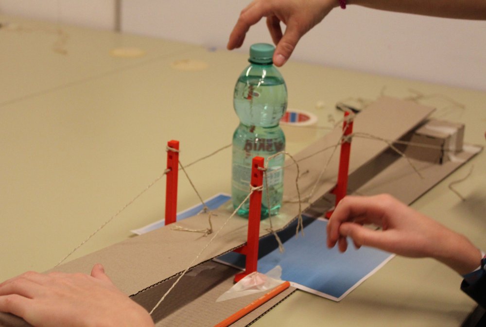 Hände, die an einer Brücke arbeiten. Auf der Brücke eine Wasserflasche.