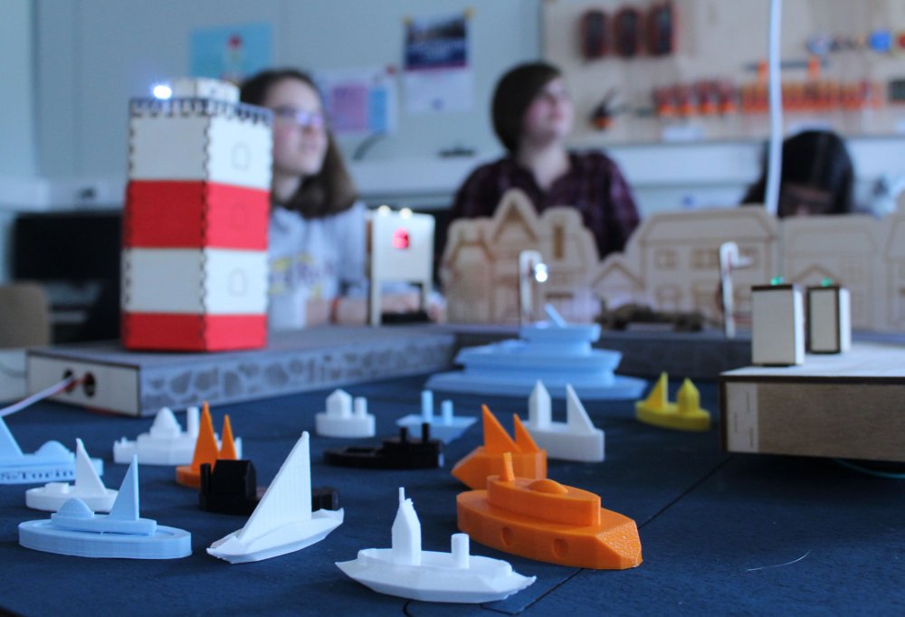 Ein Leuchtturm und Boote aus dem 3D-drucker, dahinter sidn verschwommen Menschen zu erahnen.