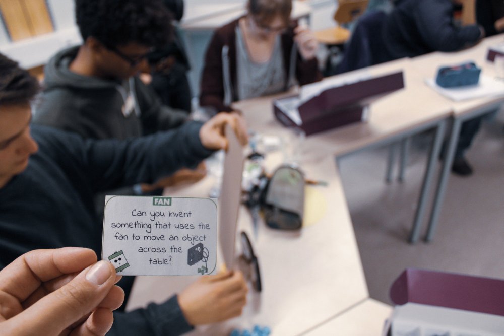 Hand häält Karte mit Erfindungsauftrag, dahinter sind verschwommen Menschen zu erkennen.