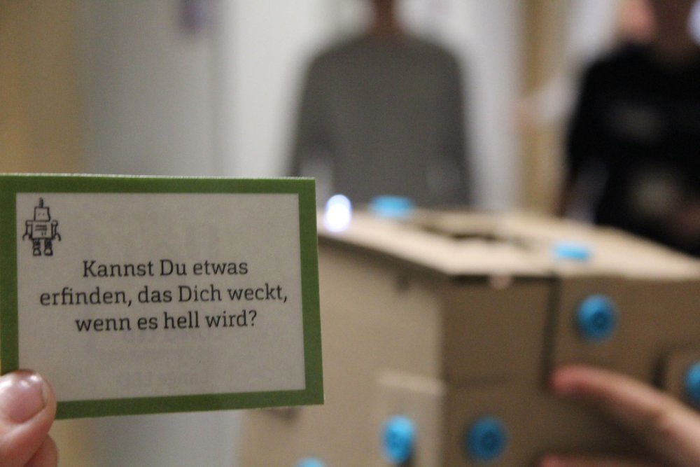 Hand häält Karte mit Erfindungsauftrag, dahinter sind verschwommen Menschen zu erkennen.