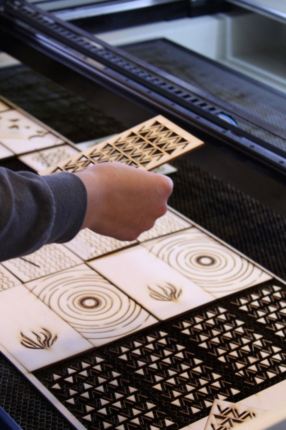 Verschiedene Muster graviert auf Holzplatten, eine Hand hält darüber ein Holzstück mit Muster.