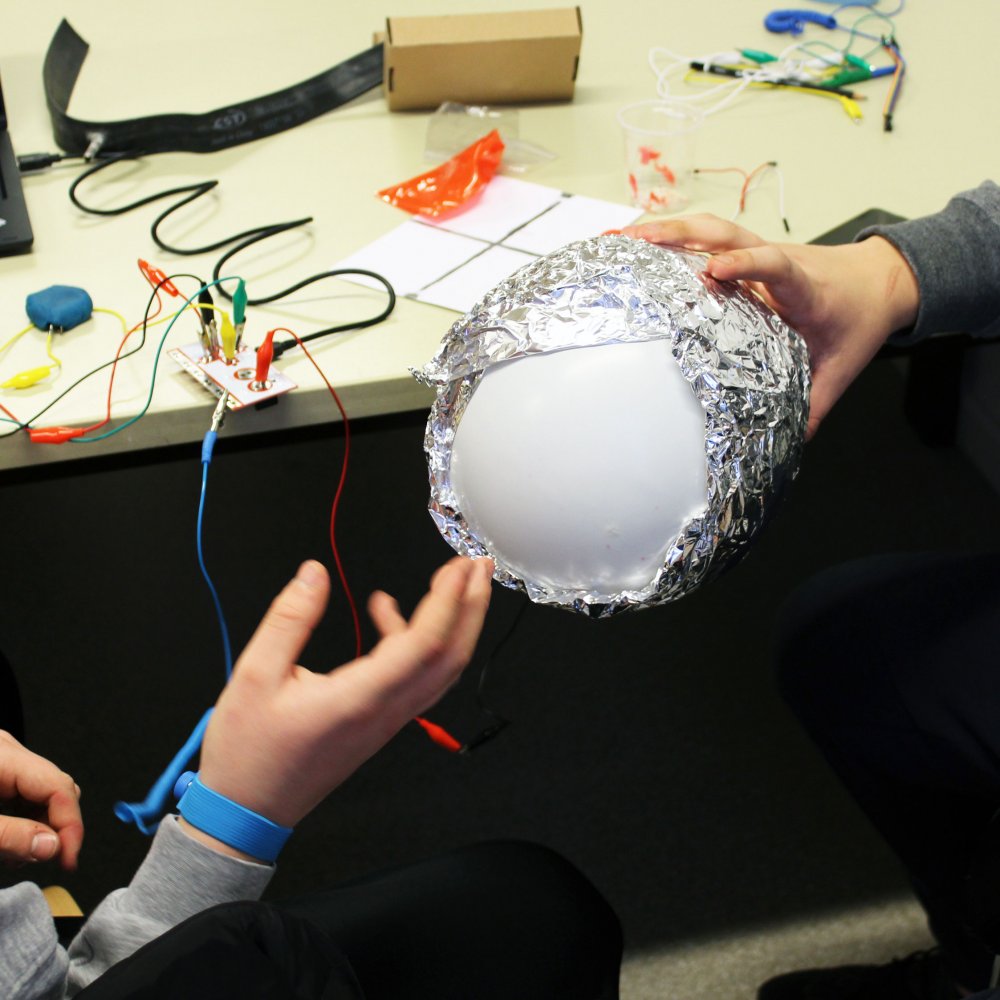 Zwei Hände, eine hält einen in Aluminium verpackten Luftballon. Auf dem Tisch dahinter kabel und andere Utensilien.