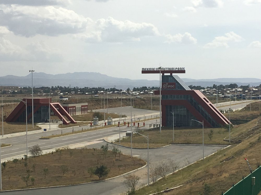 Fabrikgebäude, davor Stra0en und Wiese, dahinter bewölkter Himmel und in der Entfernung Berge.