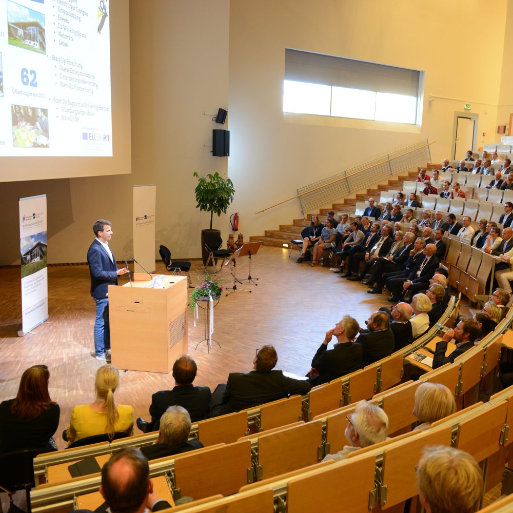Ein Saal mit vollen Sitzreihen bei einer Veranstaltung. Vorn steht ein junger Mann am Podium.