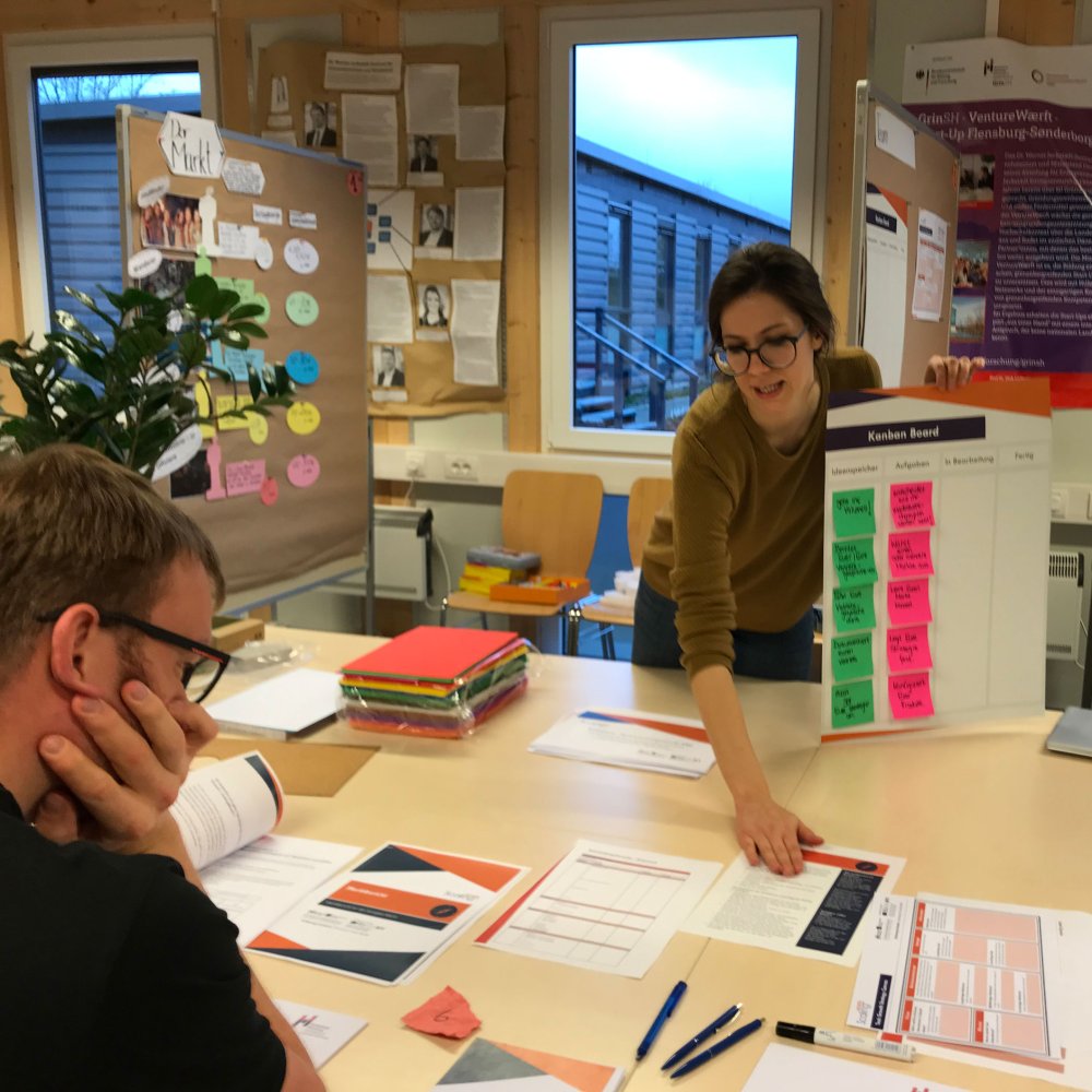 Eine Person hält einen Plan mit bunten Post-Its hoch, eine andere hört zu.