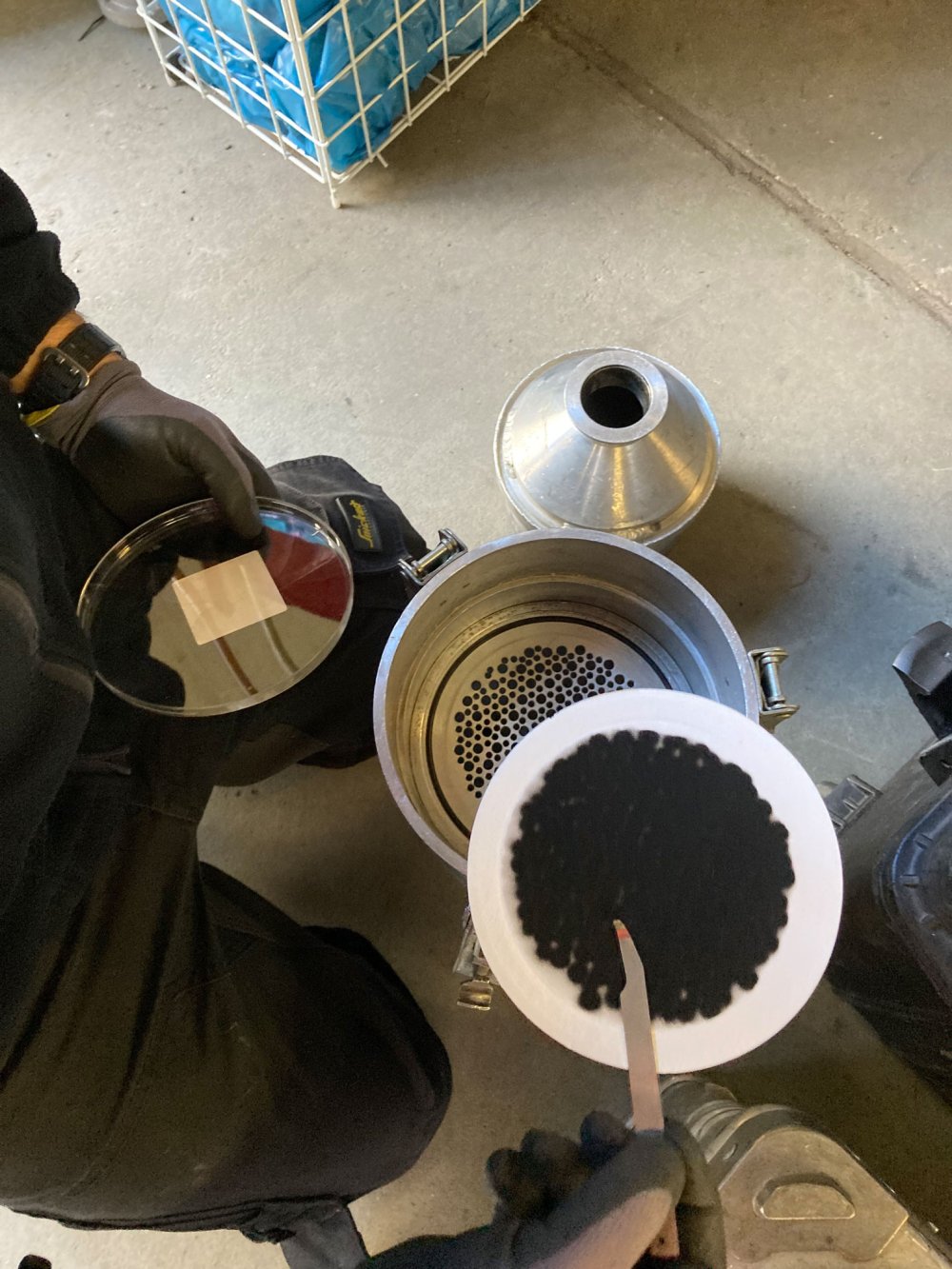 Ein Behälter mit kleinen, schwarzen Klümpchen. Daneben ein Filter. Oben eine Hand, die einen Stab hält.