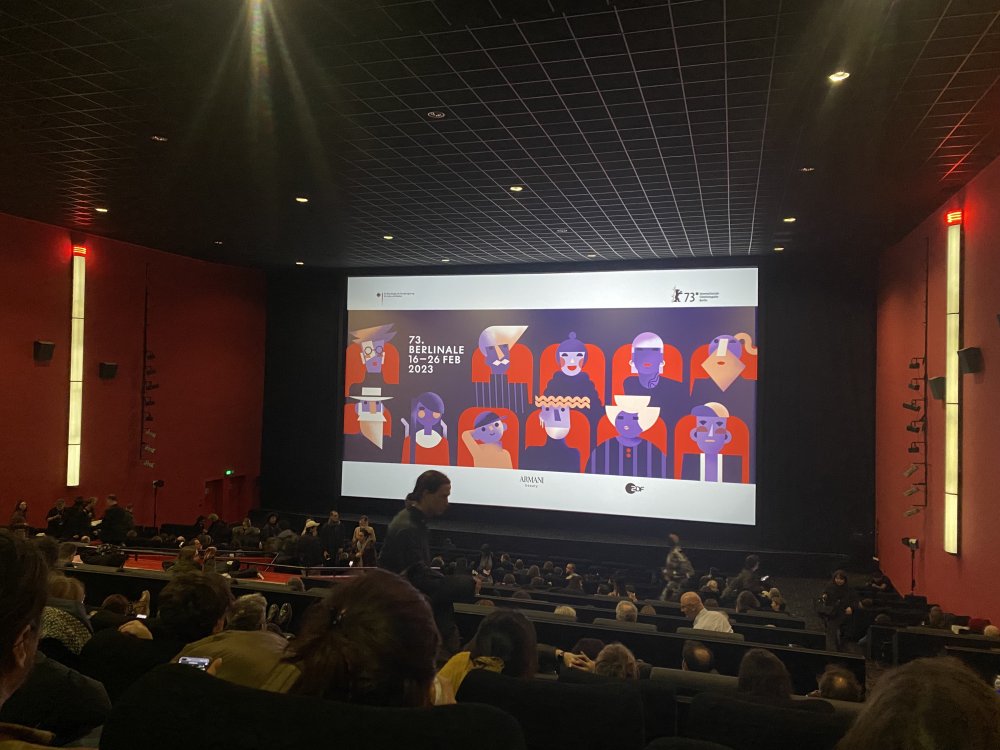 Kinosaal, vorn dunkel Publikum, dann große erleuchtete Leinwand.