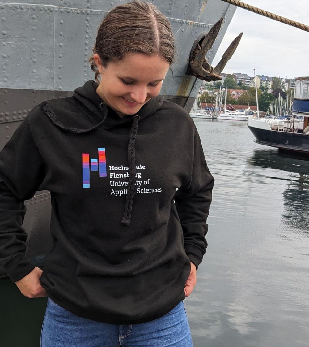Junge Frau in schwarzem Hoodie mit Hochcshuldruck steht vor einem Schiff am Flensburger Hafen.