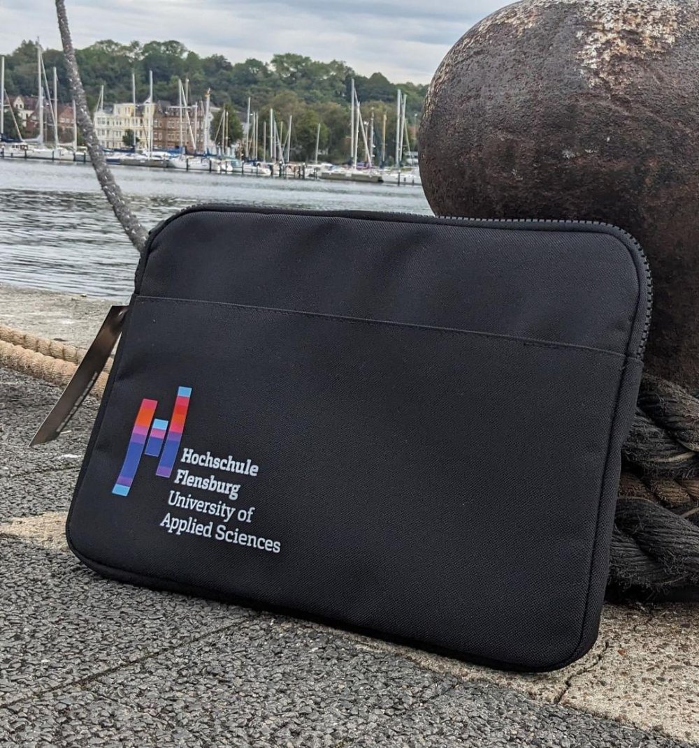 Schwarze Laptoptasche mit Hochcshuldruck lehnt an einem Stein am Flensburger Hafen.