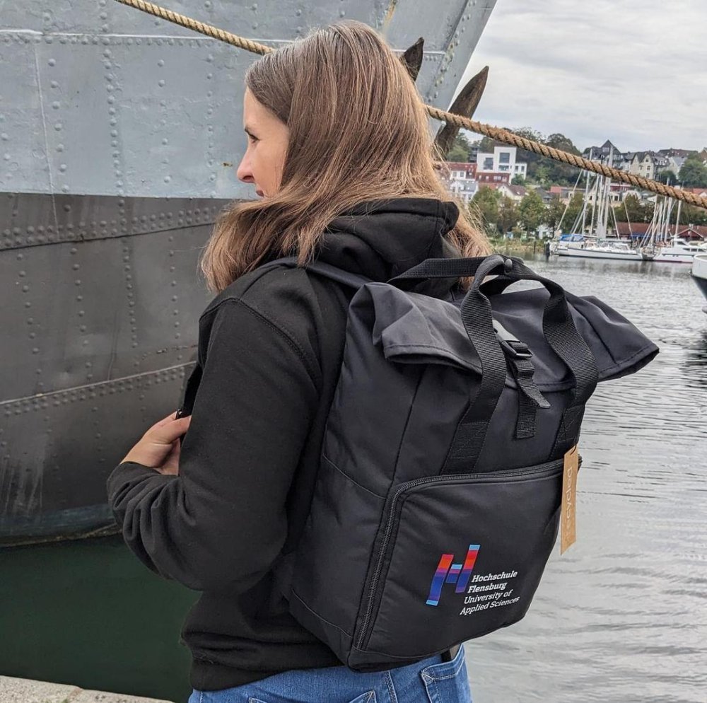 Junge Frau mit schwarzem Rucksack mit Hochcshuldruck steht vor einem Schiff am Flensburger Hafen. Sie dreht der kamera den Rücken zu.