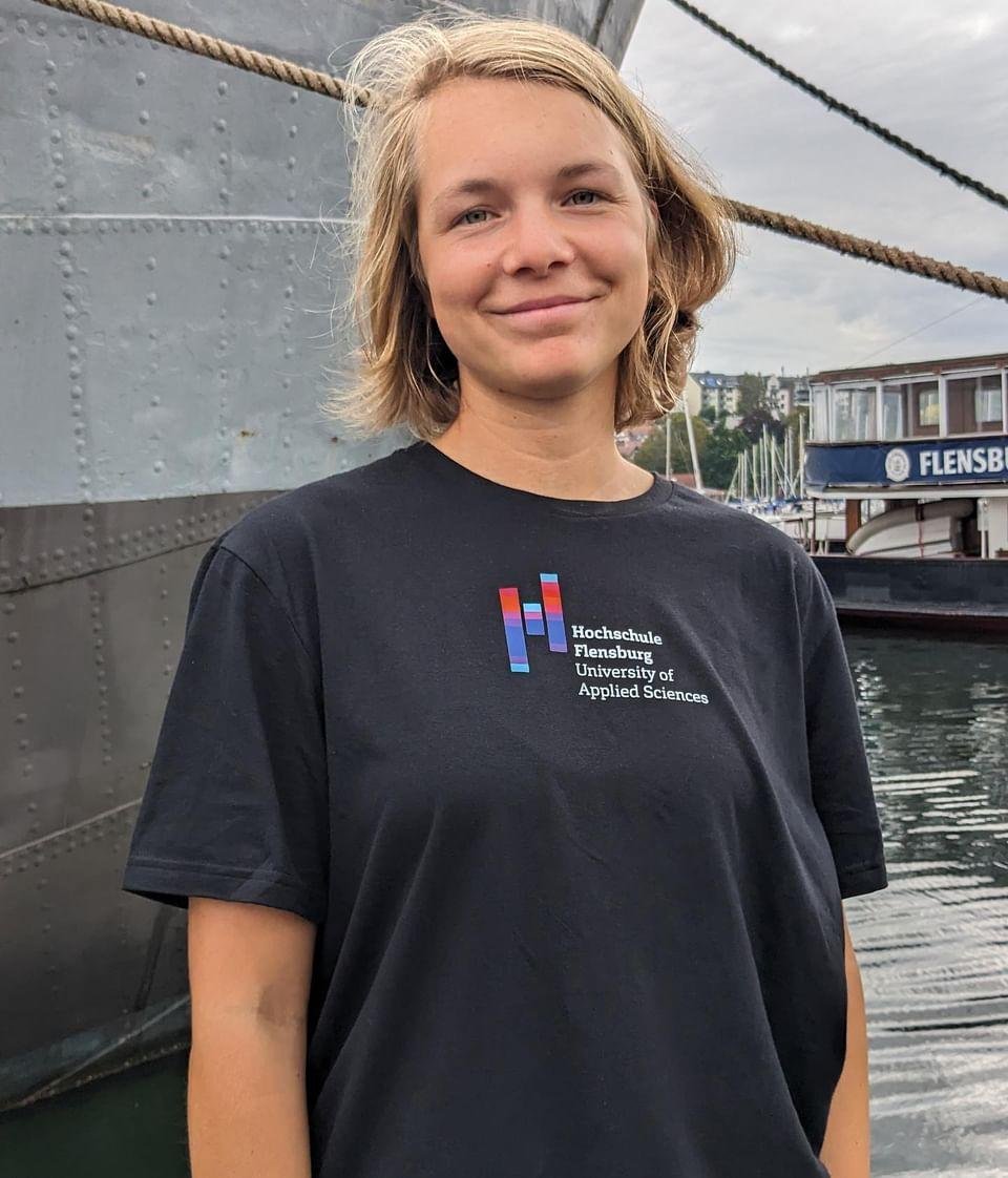 Junge Frau in schwarzem Shirt mit Hochcshuldruck steht vor einem Schiff am Flensburger Hafen. Sie lächelt in die Kamera.