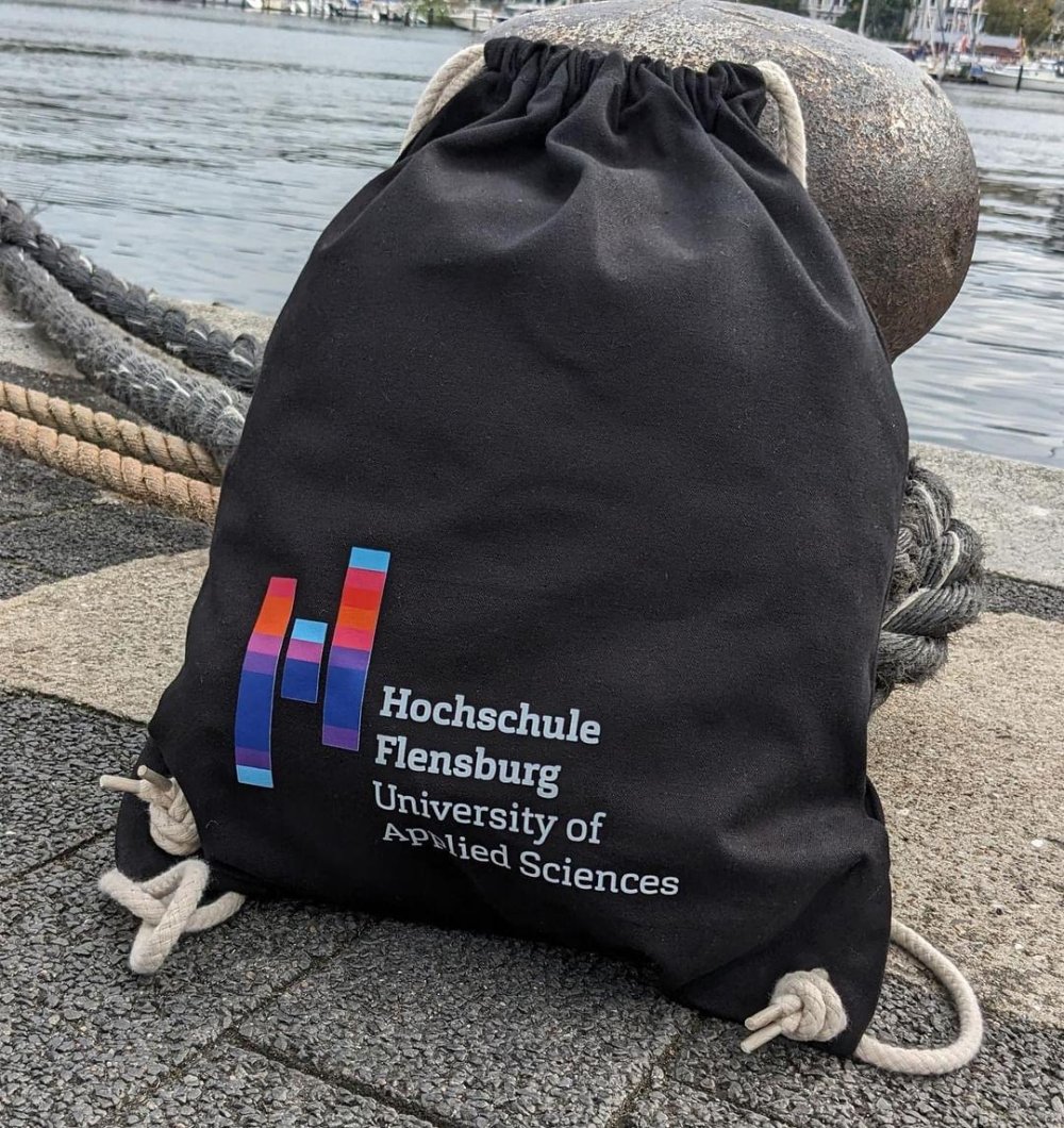 Schwarzer Turnbeutel mit Hochcshuldruck lehnt an einem Poller am Flensburger Hafen.