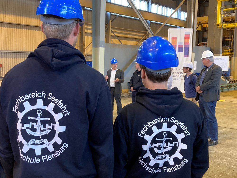 Im Vordergrund stehen zwei Studierende mit dem Rücken zur Kamera. Beide tragen Pullis mit dem Aufdruck "Fachbereich Seefahrt - Hochschule Flensburg", einem Anker und einem Zahnrad. Im Hintergrund eine Gruppe von Männern, ebenfalls mit Bauhelmen, in einer Werfthalle, und ein Aufsteller mit dem Logo der Hochschule Flensburg.