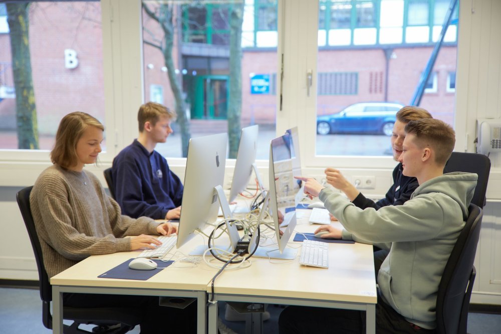Vier Studierende sitzen einnder an Schreibtischen gegenüber und schauen auf Bildschirme. Einer zeigt auf dem Bildcshirm vor ihm.