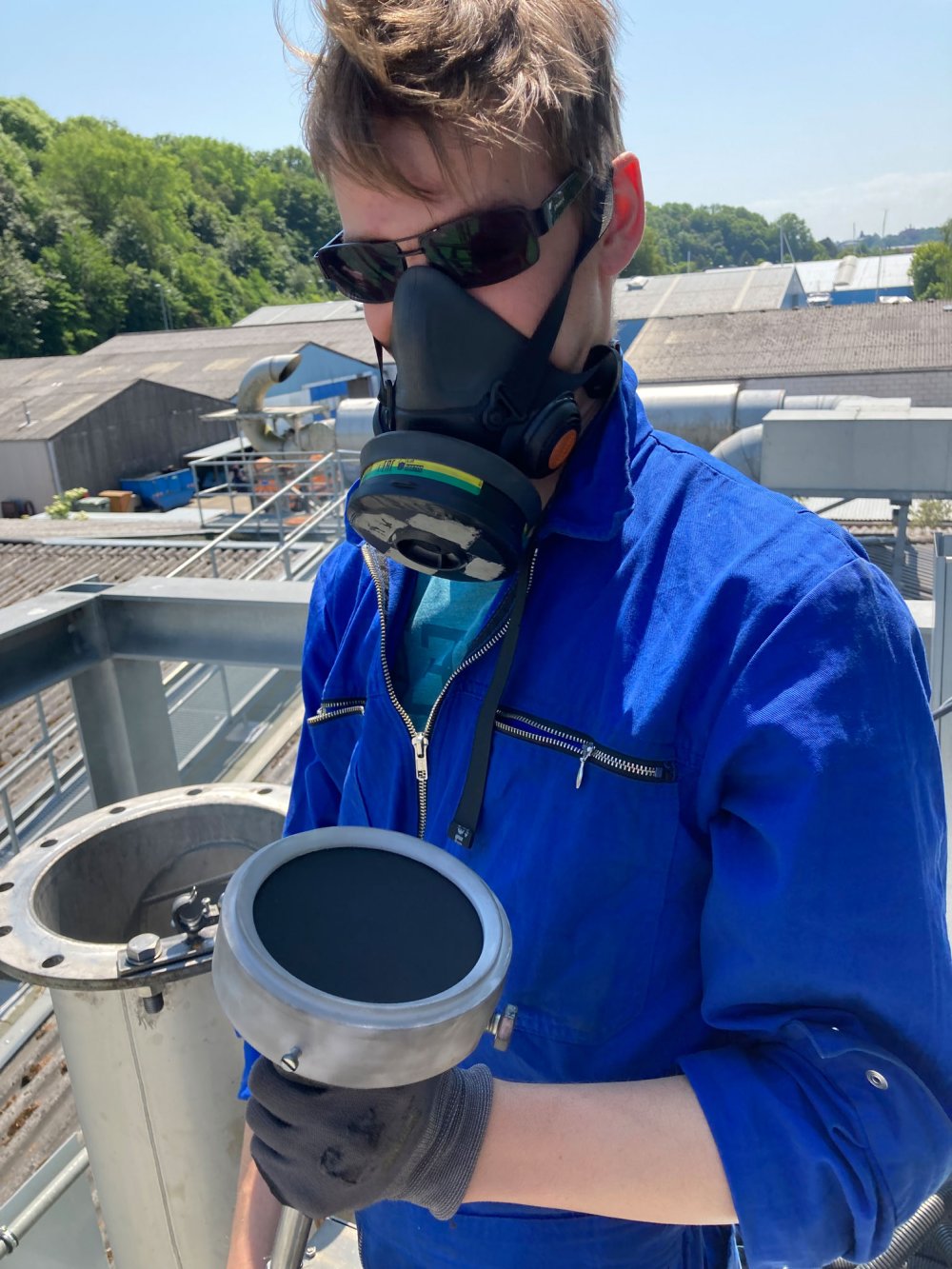 Mensch mit blauem Arbeitsanzug und Schutzmaske und -brille hält einen Filter neben einem offenen Rohr.