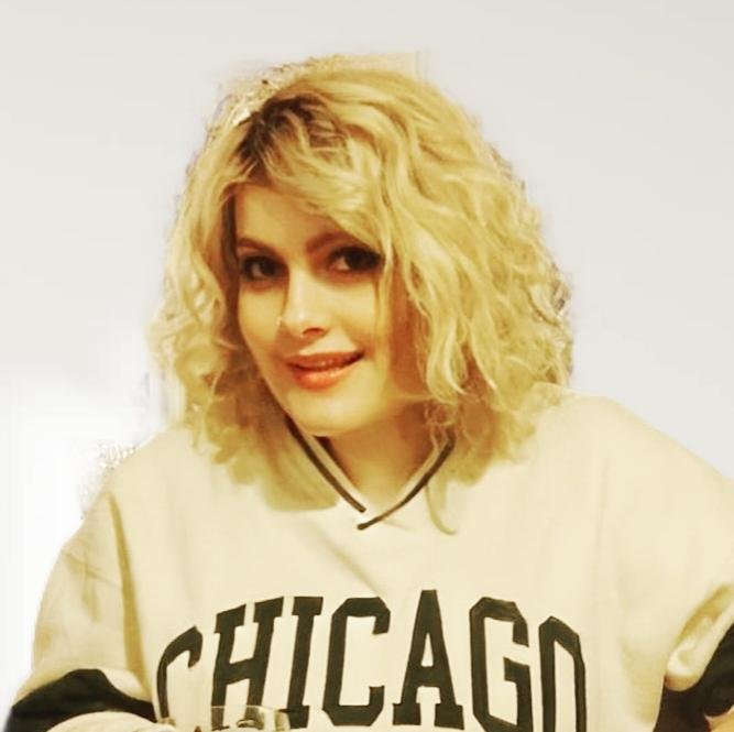 Eine junge Frau mit lockigen, blonden Haaren blickt selbstbewusst in die Kamera. Sie trägt einen weißen Pullover mit dem Aufdruck "Chicago" in Großbuchstaben.
